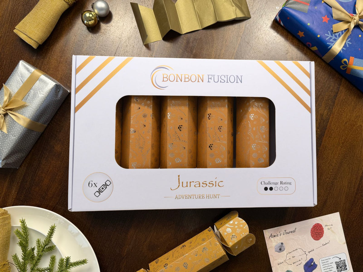Bonbon Fusion Jurassic Adventure Hunt pack of six mustard and silver foil Christmas bonbons on a festive dinner table, with a gold paper crown, a single bonbon and "Azmia's Journal" puzzle clue card