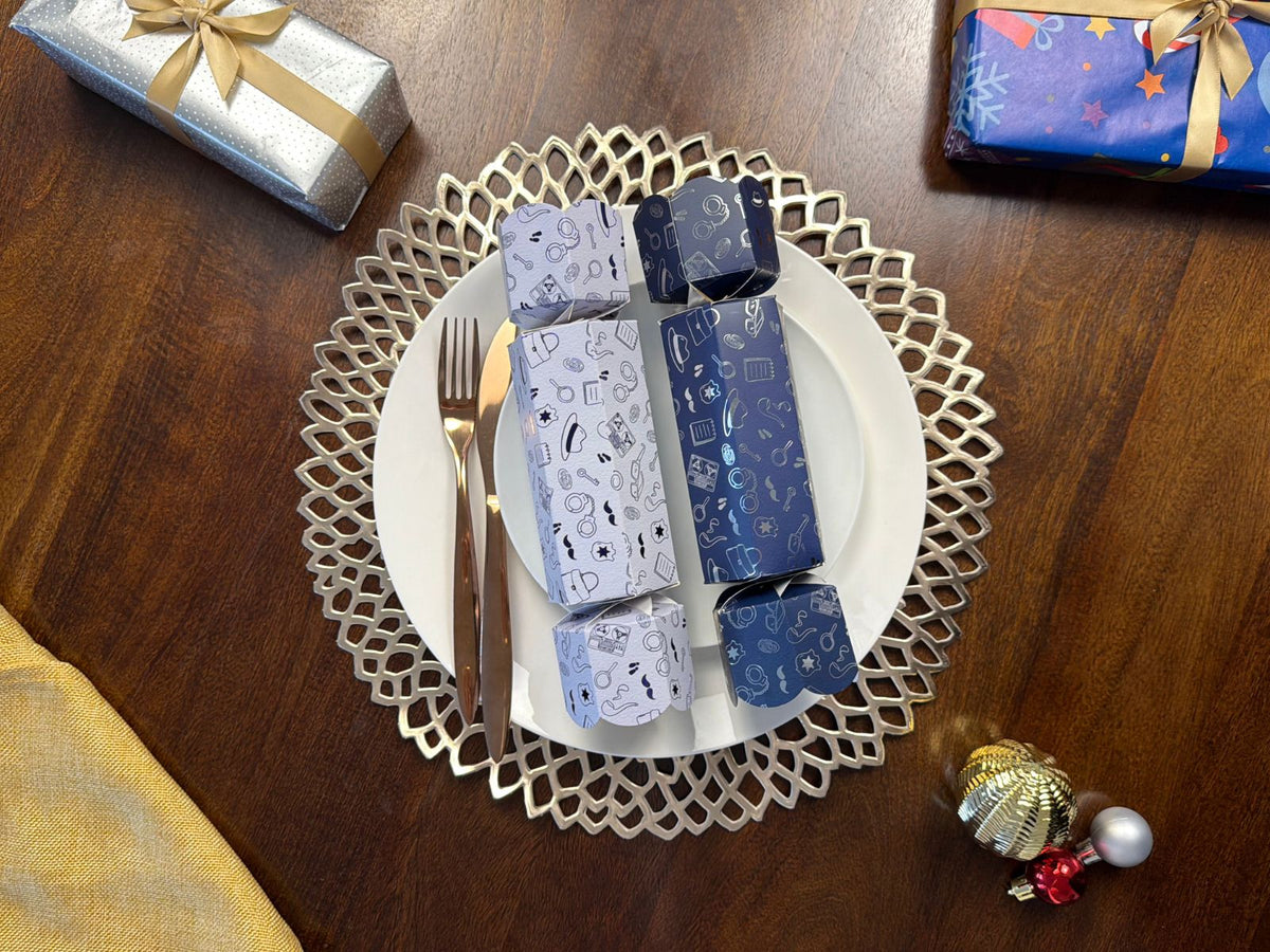 Two Bonbon Fusion The Missing Cupcake Christmas bonbons, one white and one navy with detective illustrations, on a white dinner plate with rose gold cutlery and a silver placemat on a festive Christmas table