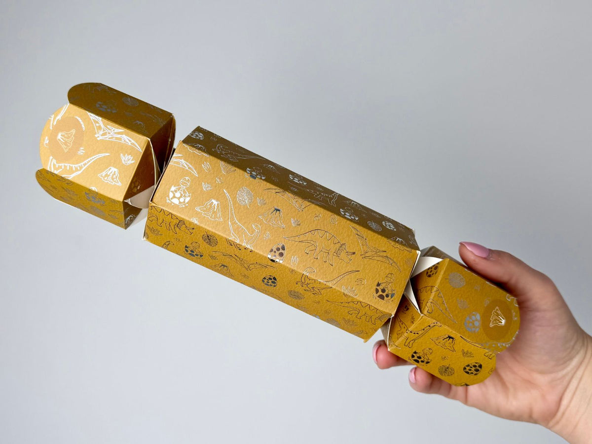 Hand holding a single Bonbon Fusion dinosaur Christmas bonbon in mustard yellow with gold and silver foil illustrations, including a triceratops, pterodactyl, dinosaur eggs, volcanoes and tropical leaves