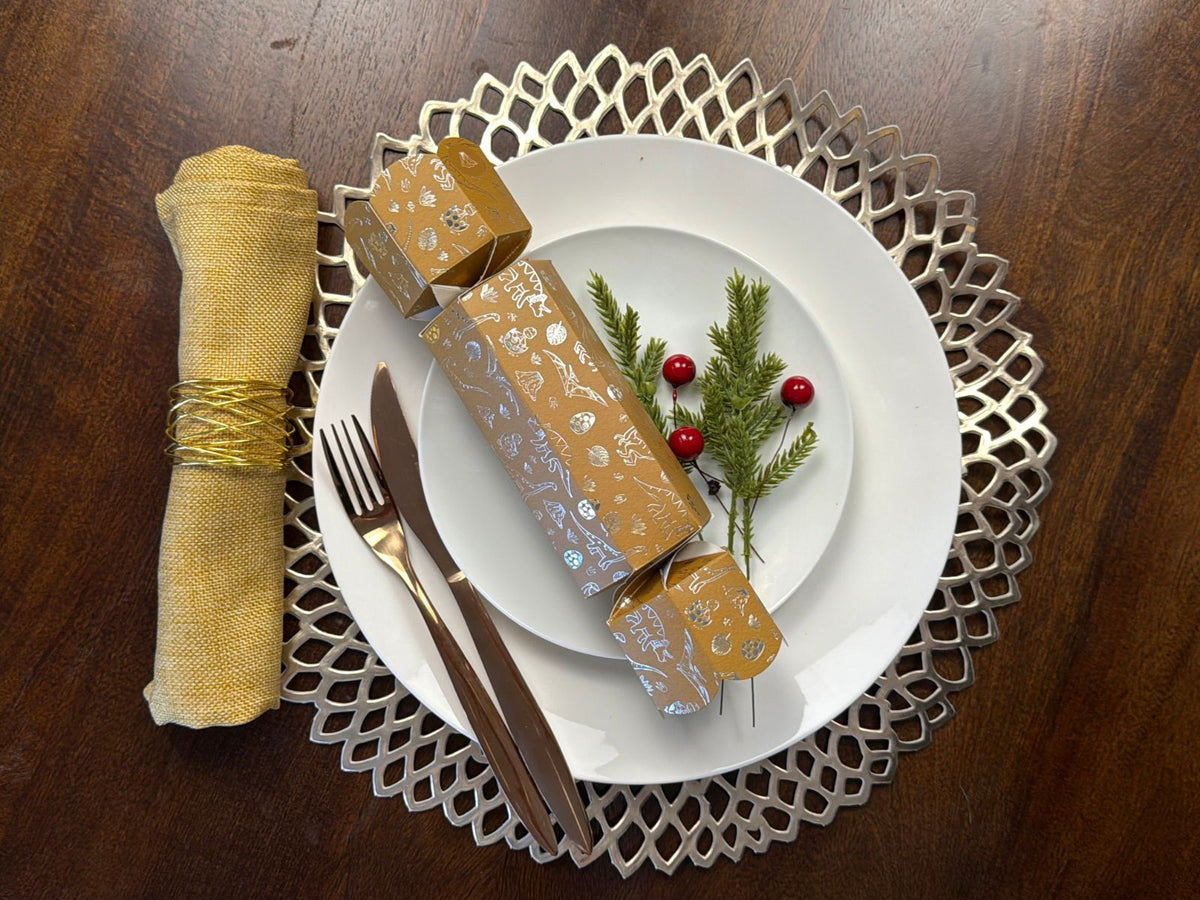 Bonbon Fusion Jurassic Christmas bonbon in mustard with silver foil dinosaur illustrations on a white dinner plate, with rose gold cutlery, a gold napkin, a silver placemat, pine sprigs and red berries on a festive Christmas table