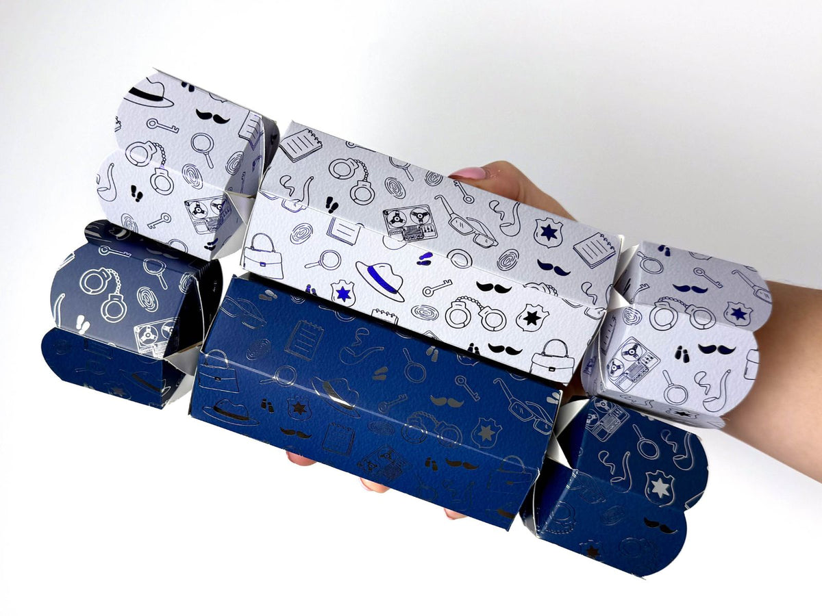 Hand holding two Bonbon Fusion The Missing Cupcake Christmas bonbons, one white and one navy with silver foil, both covered in detective illustrations including handcuffs, magnifying glasses, fingerprints, moustaches and police badges