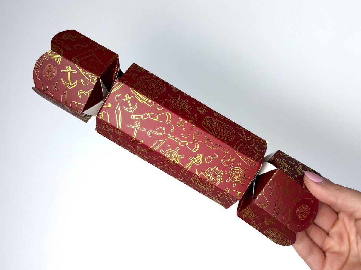 Hand holding a single Bonbon Fusion Pirates Christmas bonbon in deep red with gold foil pirate illustrations, including anchors, ship's wheels, treasure chests, pirate ships, telescopes and cutlasses
