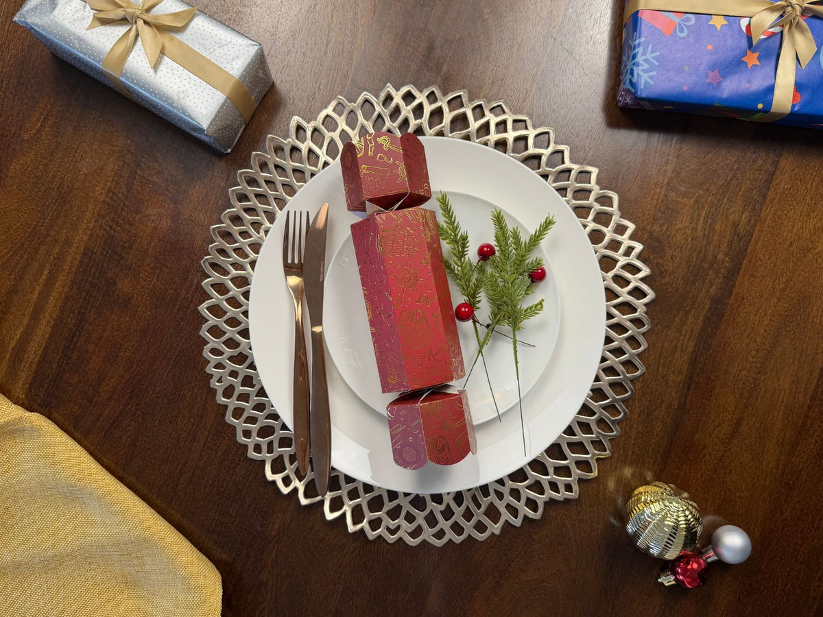 Bonbon Fusion Pirates Christmas bonbon in red with gold foil pirate illustrations on a white dinner plate, with rose gold cutlery, a silver placemat, pine sprigs and red berries on a festive Christmas table