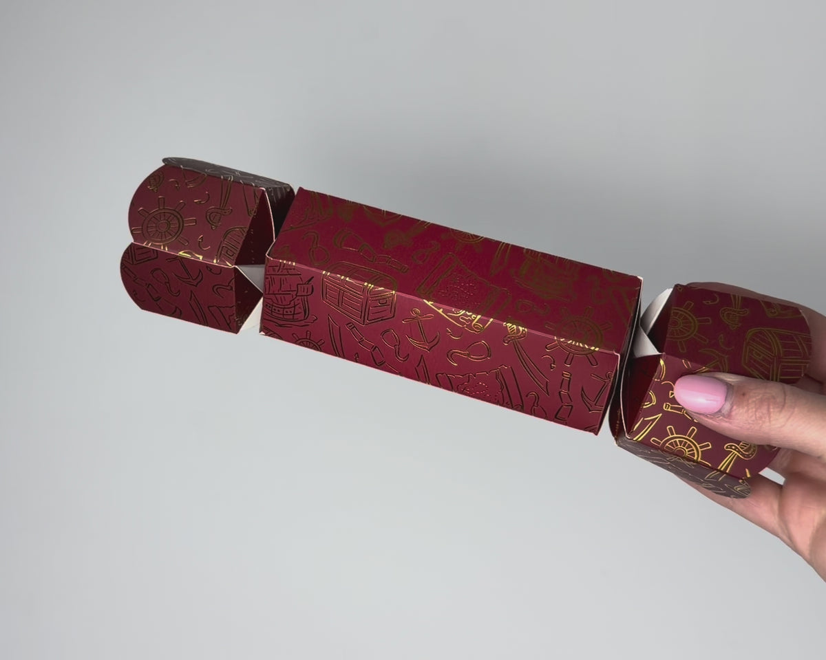 Video of a hand holding the Bonbon Fusion Pirates Christmas bonbon, showing its deep red card and shimmering gold foil pirate illustrations, including a treasure chest, pirate ship, anchors, ship's wheels and cutlasses
