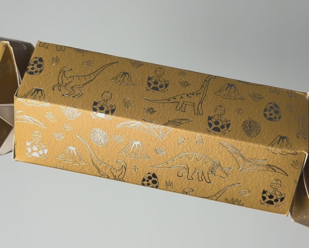 Close-up video of the Bonbon Fusion Jurassic Christmas bonbon showing its textured mustard card and shimmering silver foil dinosaur illustrations, including a triceratops, brachiosaurus, pterodactyl, baby dinosaurs hatching from eggs and volcanoes