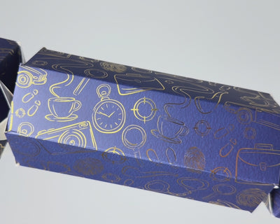 Close-up video of the Bonbon Fusion Spies Christmas bonbon showing its navy textured card and shimmering gold foil spy illustrations, including a pocket watch, teacup, camera, magnifying glass and fingerprints