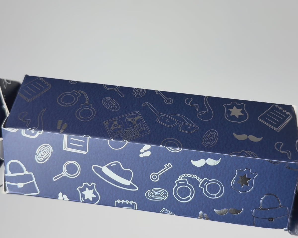 Close-up video of the Bonbon Fusion The Missing Cupcake Christmas bonbon showing its textured navy card and shimmering silver foil detective illustrations, including a fedora, handcuffs, fingerprints, magnifying glasses, moustaches and police badges