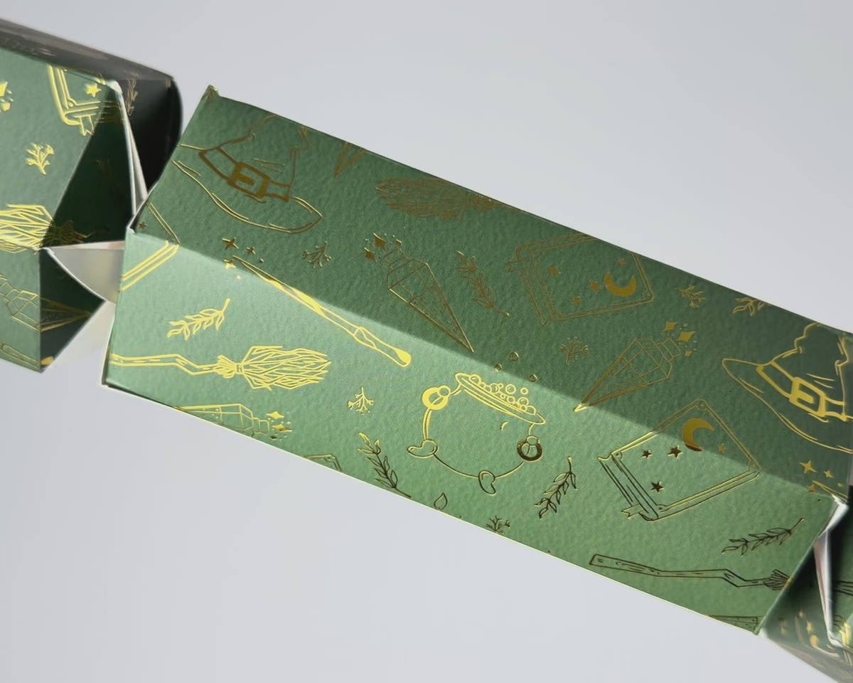 Close-up video of the Bonbon Fusion Magic Christmas bonbon showing its textured green card and shimmering gold foil magical illustrations, including a witch's hat, bubbling cauldron, broomstick, wand, potion bottles
