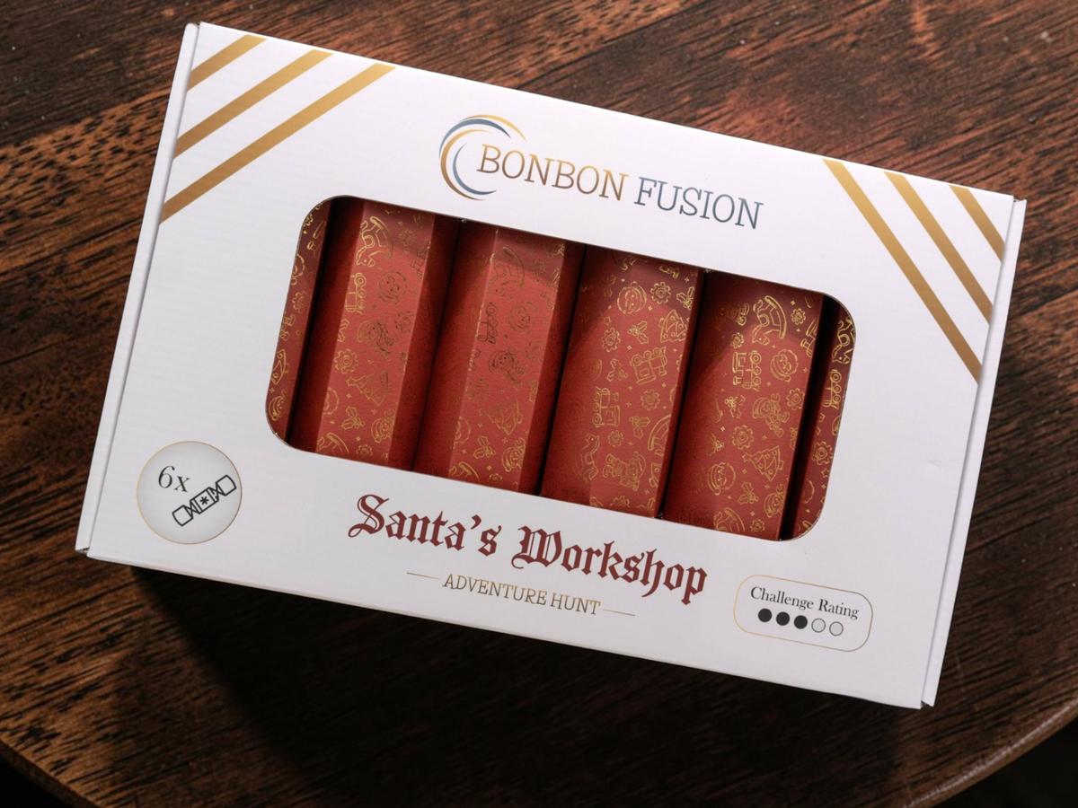 Bonbon Fusion Santa’s Workshop Adventure Hunt pack of six red Christmas bonbons with gold illustrations, in a white and gold box on a wooden table. Challenge rating: 3 out of 5.
